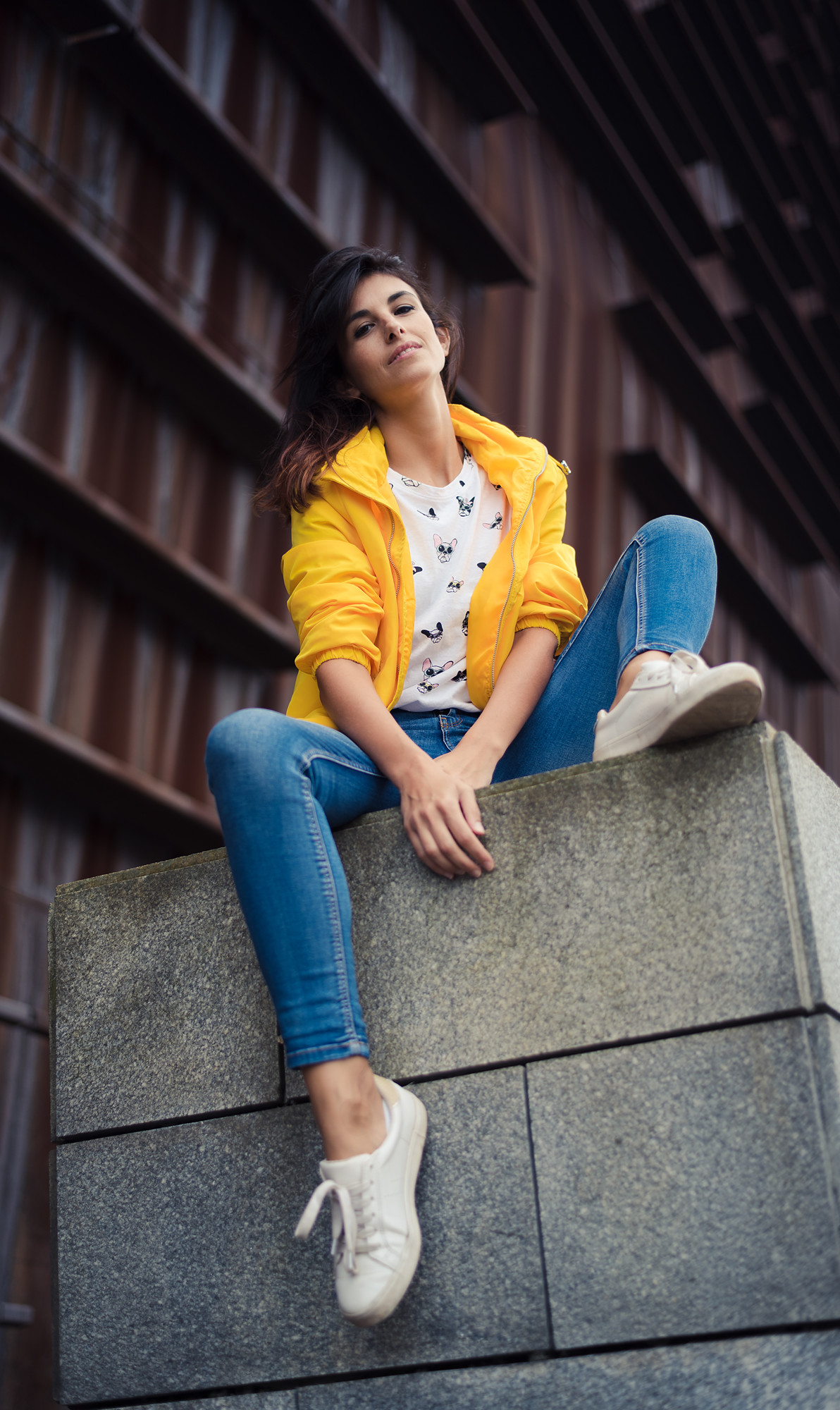Mujer morena atractiva sentada sobre un muro. Es una toma en contrapicado y ella lleva unos vaqueros y una chaqueta amarilla. Al fondo un edificio oxidado. Imagen urbana de Bilbao